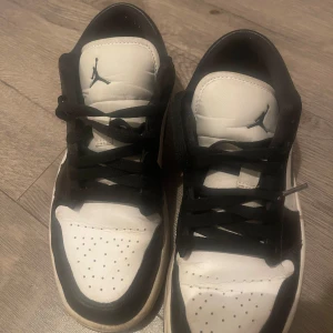 Nike Air Jordan 1 Low svart/vit - Snygga Nike Air Jordan 1 Low i svart och vitt med klassisk swoosh på sidan och Jumpman-logga på plösen. Skorna har svart snörning, perforerad tå och låg profil. Perfekta för dig som gillar streetstyle och vill ha en ikonisk sneaker i garderoben.
