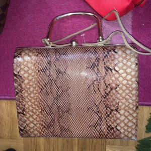 Brun handväska från Versace med ormmönster - Säljer en brun handväska från Versace med snyggt ormmönster och metallhandtag. Väskan har ett klassiskt, fyrkantigt format och detaljer i guldmetall. Märkesplakett framtill och stängs med ett elegant spänne. Perfekt för dig som gillar statement-accessoarer.