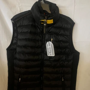 Svart dunväst från Parajumpers - Svart dunväst från Parajumpers med quiltad design, hög krage och gul detalj vid halsen. Västen har dragkedja framtill, två fickor med dragkedja och insida med orange accent. Perfekt för lager-på-lager och snygg till streetwear.