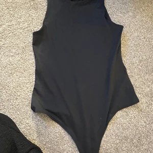 Svart ärmlös bodysuit från H&M  - Säljer en svart, ärmlös bodysuit från H&M i storlek XS. Den har hög hals och en clean, tight passform som sitter snyggt mot kroppen. Perfekt att styla med jeans eller kjol för en enkel men stilren look.