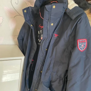 Blå parkajacka från Hampton Republic - Snygg marinblå parkajacka från Hampton Republic med fuskpäls på huvan och mysigt vitt foder. Jackan har guldiga knappar, dragkedja och coola röda detaljer samt patch på ärmen. Perfekt för kalla dagar och riktigt stilren look.