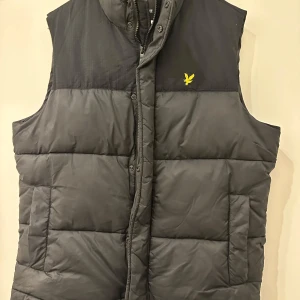 Svart dunväst från Lyle & Scott - Snygg svart dunväst från Lyle & Scott med gul logga på bröstet. Västen är quiltad och har hög krage samt dragkedja och tryckknappar framtill. Perfekt att slänga över en hoodie för extra värme. Klassisk och stilren design.