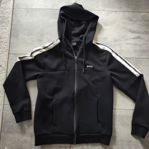 Svart hoodie från BOSS med vita ränder - Svart hoodie från BOSS med vita ränder längs axlarna och ärmarna. Tröjan har dragkedja framtill, huva med snörning och diskret BOSS-logga på bröstet. Knappt kommit till användning. (kan vara liten i storleken) 