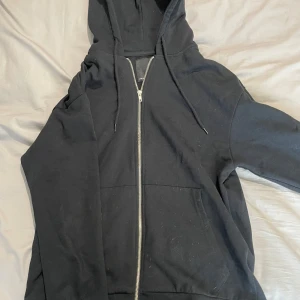 Svart hoodie med dragkedja från 157 - Svart hoodie från 157 i storlek S. Klassisk modell med huva, snörning och hel dragkedja framtill. Mjuk bomullskänsla och rymliga fickor på magen. Perfekt för chill dagar eller när du vill ha en enkel och stilren look.