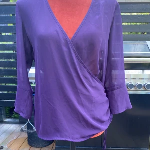 Lila omlottblus från Cappuccini - Lila blus med omlottdesign och långa ärmar. Blusen har en justerbar dragsko i sidan för en smickrande passform. Färg på bild nr 2 stämmer bäst! ﻿Längd: 66 cm ﻿polyester  Inga bilder när den sitter på mig! Katter finns i hemmet!