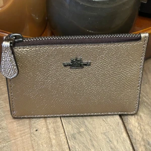 Guldig plånbok från Coach med dragkedja - Slimmad och stilren plånbok från Coach i guldigt skinn med struktur. Plånboken har dragkedja upptill, fack för kort och ett transparent ID-fönster på baksidan. Perfekt för att hålla koll på kort och cash i fickan eller väskan. ca 11 x 7,5 cm