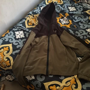 Under Armour vindjacka med huva - Snygg vindjacka från Under Armour i olivgrönt och mörkbrunt. Jackan har huva, hel dragkedja och två fickor framtill. Perfekt för blåsiga dagar och sportiga outfits. Under Armour-logga på bröstet och ärmen samt SC-detalj på ryggen.