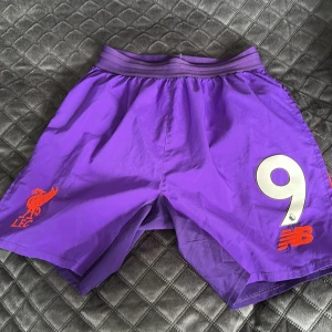 Äkta Lila Liverpool shorts New Balance - Säljer ett par lila Liverpool FC shorts från New Balance med tryckt nummer 9 och Premier League-logga. Shortsen har elastisk midja och är gjorda i lätt polyester, perfekta för fotbollsträning eller match. Klassisk sportig look med klubbmärke i rött på ena benet.