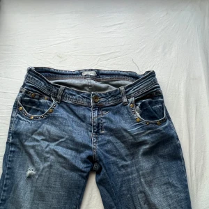 Vintage blå jeans med nitar och detaljer - Snygga blå jeans med coola nitar och dekorativa detaljer på både fram- och bakfickor. Jeansen har klassisk femficksdesign, låg midja och raka ben. Materialet är jeans i bomull och de har slitningar de är i storlek 31/34.