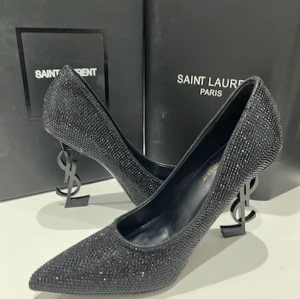 Svarta Saint Laurent pumps med strass - Exklusiva svarta pumps från Saint Laurent, täckta med glittrande strass och med ikonisk YSL-logga som klack. Skorna har spetsig tå och hög klack, perfekt för dig som vill sticka ut. Materialet är skinn och syntet, och designen är både lyxig och edgy.