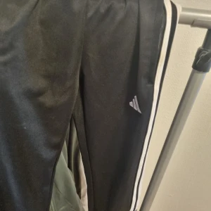 Svarta Adidas mjukisbyxor med vita ränder - Svarta mjukisbyxor från Adidas med klassiska vita ränder längs sidorna och Adidas-logga på benet. Byxorna har resår i midjan och är gjorda i ett mjukt, syntetmaterial som passar perfekt för träning eller chill. Snygg och sportig look.