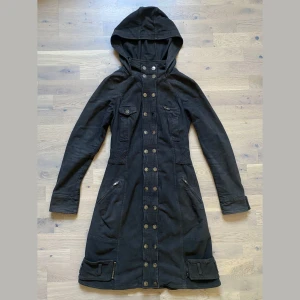 Gaetano Navarra F/W 2004 Brown Button-Up Dress Coat - Gaetano Navarra brun klänning/kappa med huva, ribbad midja, nederkantsbälte och knäppning framtill. 97 % bomull, 3 % elastan. Tillverkad i Italien. På etiketten står det storlek 40, men passar som en 36. Bröstvidd 41,5 cm, midja 36,5 cm, längd 92 cm, ärmlängd 65 cm.