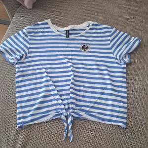 Randig blåvit knyt-topp från Cropp - Superfin blåvit randig topp från Cropp med korta ärmar och knytning framtill. T-shirten har ett marin-inspirerat märke med ankare på bröstet och är perfekt för en chill sommarlook.