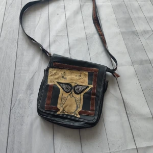 Svart axelväska med uggla och tidningsprint - You can buy this only on Vinted profile: brysu1 Cool svart axelväska i skinnimitation med bruna detaljer och unik uggla framtill, dekorerad med prickiga ögon och tidningsinspirerat print. Väskan har flera fack, dragkedja och justerbar axelrem. Perfekt för dig som vill sticka ut med din stil. 