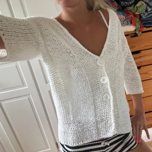 Vit stickad kofta från Oui - Vit stickad kofta från Oui med trekvartsärm och stora vita knappar framtill. Koftan har en djup v-ringning och är gjord i ett grovstickat material som känns luftigt och skönt. 