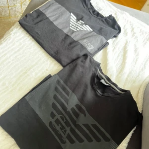 Svarta EA7 Emporio Armani t-shirts - Två svarta t-shirts från EA7 Emporio Armani med stora logotryck framtill. Ena t-shirten har en grå panel över bröstet och båda har klassisk rund hals. Snygg och stilren design som passar perfekt till jeans eller shorts.