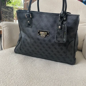 Snygg svart handväska från Guess - Den har logomönster över hela väskan och har silverfärgade metalldetaljer. Dubbla handtag och flera fack med dragkedja. Den är i mycket bra skick ser helt ny ut. Köpt över 2000 kr men säljer för 850. Priset kan diskuteras!❤️och den är även perfekt till att resa