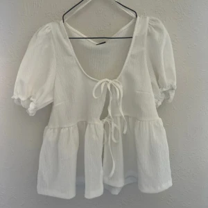 Vit blus med puffärm och knyt - Superfin vit blus med puffiga korta ärmar och två knyt framtill. Blusen har en luftig och flowy passform med volanger nedtill och djup urringning. Perfekt för en somrig look. Aldrig använd.