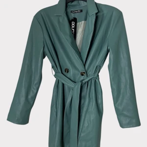 Grön skinn trenchcoat från Boohoo - Trendigt grön trenchcoat i skinnimitation från Boohoo. Jackan har långa ärmar, knytskärp i midjan och dubbla knappar framtill. Snygg krage och stilren design som ger en cool vibe. Perfekt för dig som vill sticka ut med en unik jacka. (Passar mig som har storlek 36-38) 