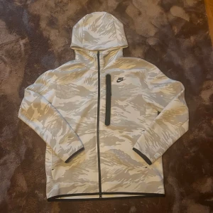 Nike camo vindjacka med huva - Nike tröja  i beige och grått camomönster med hel dragkedja och huva. Tröjan har en svart bröstficka med dragkedja och Nike-logga på bröstet. Perfekt för dig som gillar sportig och street-inspirerad stil.