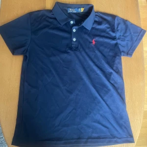 Ralph Lauren Piké - Stilren Ralph Lauren Piké i färgen mörkblå med röd logga, i ett träningsliknande material.  Den är i princip nyskick förutom att etiketten sitter löst i ena änden. (bild 3). Bara att skriva vid någon fråga 🙌