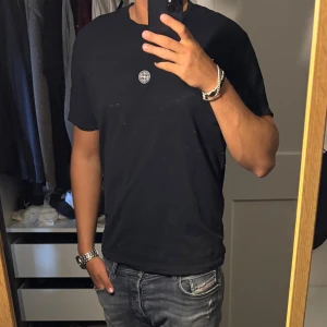 Svart Stone Island t-shirt i bomull - Svart t-shirt från Stone Island med klassisk logga på bröstet och stor vit Stone Island-tryck på ryggen. Skön passform och rund halsringning. Tillverkad i mjuk bomull, perfekt för en clean och stilren look.