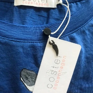 Blå t-shirt Coster Copenhagen snake - Blå t-shirt från Coster Copenhagen med tryck av en rosa och svart orm och texten 'Pink as Love' på bröstet. T-shirten har korta ärmar och rund hals. Materialet är mjuk bomull, perfekt för en cool och avslappnad stil.