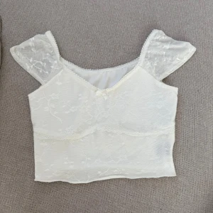 Vit topp - Säljer en söt vit croppad topp med transparenta spetsärmar och diskret blommönster. Toppen har fina spetsdetaljer längs kanterna och är perfekt för en somrig look.