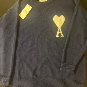 Mörkblå stickad tröja  - Snygg mörkblå stickad tröja med ikoniskt hjärta och A-logga i vitt på bröstet. Tröjan har rund hals, ribbade muddar och är långärmad. Perfekt för dig som gillar stilrena och trendiga plagg med en twist. OBS!!! INTE ÄKTA