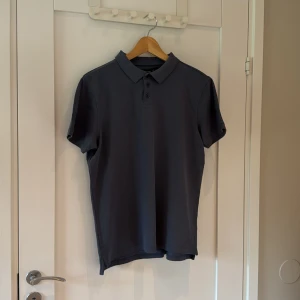 Mörkgrå pikétröja från 157 - Snygg mörkgrå pikétröja från 157 i storlek L. Klassisk krage och tre knappar framtill. Tillverkad i bomull med en lätt strukturerad yta. Perfekt för en clean och stilren look. Kortärmad modell som passar till många outfits.