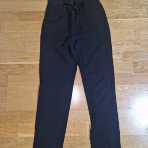 Svarta byxor med knyt i midjan - Svarta byxor med hög midja och knytband, elastisk midja och raka ben. Byxorna har fickor fram och bak, samt bälteshällor. Perfekta för en chill och stilren look. Materialet känns mjukt och bekvämt, passar till många outfits.