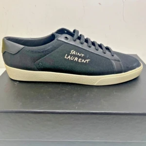 Svarta sneakers från Saint Laurent - Stilrena svarta sneakers från Saint Laurent med vit sula och handskriven logga på sidan. Skon har snörning och är tillverkad i canvas med detaljer i skinn. Perfekt för dig som gillar minimalistisk och lyxig streetstyle.