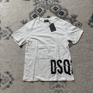 Vit DSQUARED2 t-shirt med tryck - Vit t-shirt från DSQUARED2 med svart DSQ-tryck nere vid fållen. Klassisk rund hals och korta ärmar. Tillverkad i mjuk bomull för en skön känsla och clean look. Perfekt för dig som gillar stilrena plagg med en twist.