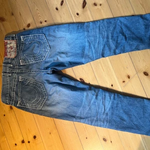 True Religion jeans blå - Säljer ett par klassiska blå jeans från True Religion med tydliga vita kontrastsömmar och ikoniska bakfickor. Jeansen har rak passform och normal midja. Tillverkade i USA av slitstarkt bomullsjeans. De är använda men riktigt feta! De sitter som en smäck👌 Hör av er vid frågor!🙏