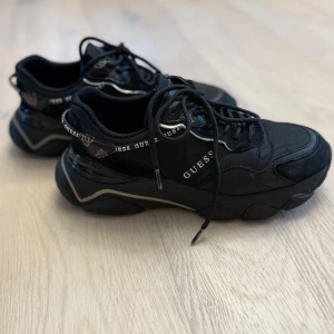 Svarta chunky sneakers från Guess - Svarta chunky sneakers från Guess med logga längs sidorna och detaljer i guld. Skorna har snörning, rund tå och en kraftig sula. Materialmix av textil och syntet med svarta och guldiga detaljer som ger en cool och sportig vibe.