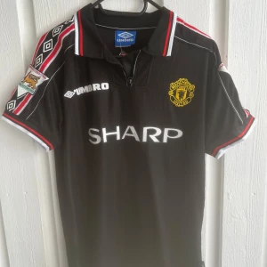 Manchester United Beckham tröja Umbro - Klassisk svart Manchester United fotbollströja från Umbro med röda och vita detaljer. Nummer 7 och Beckham på ryggen, SHARP-logga på bröstet och klubbmärke i gult. Snygga mönster på ärmarna och krage med röd och vit kant. Perfekt för fans av 90-talsfotboll.
