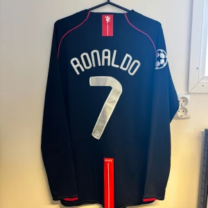 Manchester United Ronaldo 7 långärmad tröja - Snygg svart långärmad Manchester United-tröja från Nike med Ronaldo 7 på ryggen. Champions League-märke på ärmen, röda detaljer och AIG som sponsor på bröstet. Perfekt för dig som älskar fotboll och vill sticka ut med en ikonisk supportertröja.
