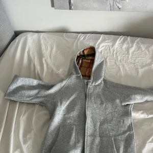 Grå hoodie från Burberry med dragkedja - Snygg grå hoodie från Burberry med klassiskt rutigt foder i huvan och broderad logga på bröstet. Det står XL på storlekslappen för den är XL i barnstorlekar som är S/M i vanligt. Tröjan har dragkedja framtill, kängurufickor och är tillverkad i mjuk bomull. Perfekt för en chill och stilren look. 