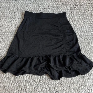 Svart volangkjol i bomull - Svart kortkjol med volangkant längst ner. Kjolen har en lätt rynkad detalj på ena sidan och är gjord i mjuk bomull. Perekt för en sommardag eller en utekväll. Aldrig använd. Går att vika ner om man vill ha den lågmidjad.