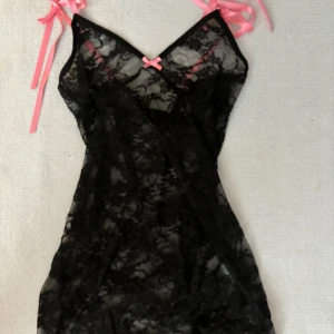 Svart spetsklänning med rosa rosetter - Snygg kort svart spetsklänning med transparent material och rosa rosetter vid axelbanden och bysten. Axelbandslös design med knytband i rosa för en söt kontrast. Perfekt för dig som gillar en lekfull och feminin stil.