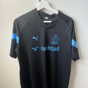 Marseille svart matchtröja Puma - Snygg svart fotbollströja från Puma med Olympique de Marseille-logga och sponsortryck i ljusblått. Tröjan har korta ärmar med ljusblå detaljer och är tillverkad i ett lätt och ventilerande material som passar perfekt till träning eller match.