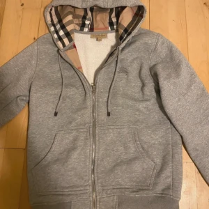 Grå hoodie från Burberry med dragkedja - Grå hoodie från Burberry med klassiskt rutigt foder i huvan och broderad logga på bröstet. Tröjan har dragkedja, snörning i huvan och stora fickor framtill. Tillverkad i mjuk bomull med elastan för extra komfort.