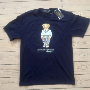 Mörkblå Polo Bear t-shirt Ralph Lauren - Snygg mörkblå t-shirt från Ralph Lauren med ikoniska Polo Bear-trycket på bröstet. Klassisk rund hals och korta ärmar. Tillverkad i mjuk bomull som känns skön mot huden. 
