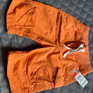 Orange shorts med snörning från KappAhl - Snygga orange shorts från KappAhl med vit snörning i midjan och bred resår. Shortsen har praktiska fickor på sidorna och är gjorda i mjuk bomull, perfekta för varma sommardagar. Enkel och avslappnad stil som passar till allt.