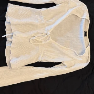 Vit topp från Gina Tricot - Supersöt vit topp från Gina Tricot i storlek S. Toppen har långa ärmar, v-ringning och knyt framtill. Materialet är lätt och strukturerat med en vågig känsla, perfekt för en fräsch och trendig look.