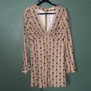 Mönstrad kortklänning från Pull&Bear - Säljer en retroinspirerad kortklänning från Pull&Bear med lång ärm och djup v-ringning. Klänningen har ett färgstarkt mönster i lila, orange, svart och grön samt dragkedja bak. Perfekt för dig som gillar unika prints och coola vibbar. Använda 1 gång