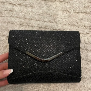 Svart glittrig kuvertväska med kedja - Snygg svart kuvertväska med glittrig yta och silvrig metallkedja. Väskan har ett elegant V-format metallspänne framtill och magnetlås. Perfekt storlek för mobilen och det viktigaste. Insidan är fodrad i svart tyg och kedjan kan tas av. Använd 1 gång