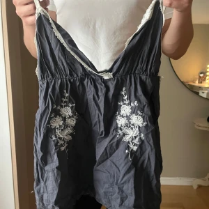 Mörkblå linnetopp med spets och broderi - Supersöt mörkblå linnetopp med tunna axelband, vit spetskant och vita blommiga broderier framtill. Toppen har v-ringning och är perfekt för en chill sommarlook. Lätt och luftig känsla med fina detaljer som gör den unik.