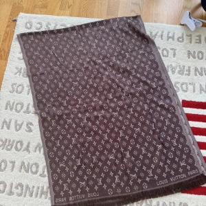 Brun Louis Vuitton halsduk med monogram - Snygg brun halsduk från Louis Vuitton med klassiskt monogrammönster och fransad kant. Stor och lyxig accessoar som lyfter vilken outfit som helst. Perfekt för dig som gillar exklusiva detaljer och vill sticka ut.