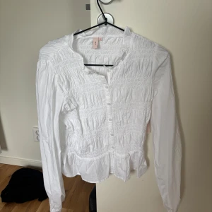 Vit blus nelly  - Säljer en vit långärmad skjorta från nelly med smockade detaljer över hela bröstet och midjan. Skjortan har knappar framtill och en lätt volang nertill. Perfekt för dig som gillar romantisk och feminin stil. vit blus från nelly 